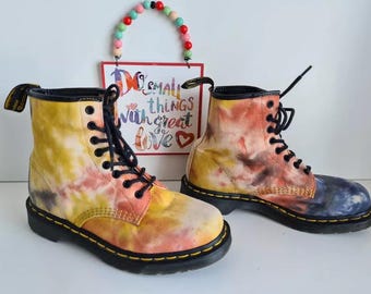 May include: A pair of tie-dye Dr. Martens boots with yellow, orange, and blue accents. The boots have black laces and a black sole.