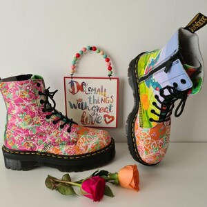 May include: A pair of colorful patterned Dr. Martens boots. The boots have black laces and a black sole. The boots are decorated with a variety of colors and patterns, including pink, blue, green, and yellow. The boots are sitting on a white surface. The text "Do small things with great love" is visible in the background.