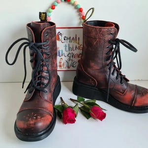 Könnte beinhalten: Ein Paar rote Schnürstiefel aus Leder mit schwarzen Schnürsenkeln und einer dicken schwarzen Sohle. Die Stiefel haben einen abgenutzten Look und sind getragen. Die Stiefel stehen auf einer weißen Oberfläche mit einem kleinen Schild, auf dem "Kleine Dinge mit großer Liebe" steht.