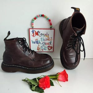 Può includere: Un paio di stivali da donna in pelle marrone con lacci neri. Gli stivali hanno una suola spessa in gomma nera. Gli stivali sono su una superficie bianca con un cartello che dice "Do small things with great love".