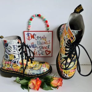 May include: A pair of black and white Dr. Martens boots with a colorful graphic print. The boots have laces and a black sole. The boots are sitting on a white surface with a sign that says "Do small things with great love" and a heart.