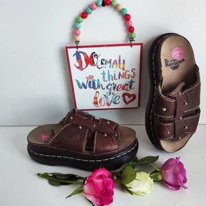 Può includere: Sandali in pelle marrone con un design a borchie e una suola spessa a piattaforma. I sandali hanno un'etichetta del marchio Dr. Martens sul cinturino. I sandali sono su una superficie bianca con un cartello che dice "Do small things with great love" e una collana di perline colorate.