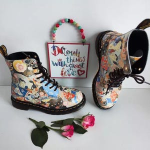 Könnte beinhalten: Paar schwarze und mehrfarbige Stiefeletten mit einem einzigartigen Muster. Die Stiefel haben einen Schnürverschluss und sind mit dekorativen Nieten verziert. Ein Schild mit dem Text "Do small things with great love" ist im Hintergrund zu sehen.