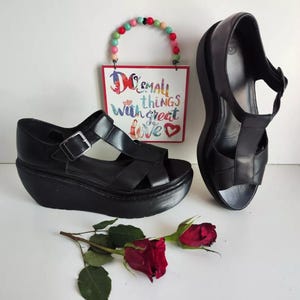 May include: Two pairs of black leather sandals. One pair has a thick platform sole and a buckle fastening. The other pair has a thin strap and a buckle fastening. The sandals are on a white surface with a sign that reads "Do small things with great love."