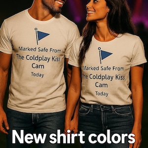 May include: Two people wearing white t-shirts with a blue flag graphic and the text "Marked Safe From The Coldplay Kiss Cam Today." They are holding hands. The text "New shirt colors now available!" is at the bottom.