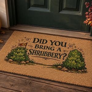 Può includere: Uno zerbino rettangolare in fibra di cocco beige con il testo nero DID YOU BRING A SHRUBBERY? centrato tra due cespugli verdi illustrati. Il design include piccole stelle decorative e una piccola icona a forma di bandiera sopra il cespuglio sinistro.