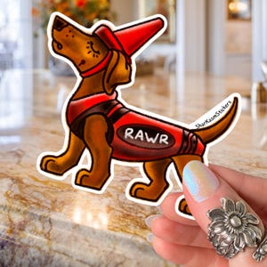 Puede incluir: Una pegatina con un perro salchicha de dibujos animados que lleva un sombrero y un chaleco de bombero rojos con la palabra "RAWR". El perro es marrón con detalles en negro y un contorno blanco. La pegatina está sujeta por una mano.