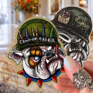 Peut inclure: Deux autocollants avec des illustrations de bouledogues. L'un porte un casque militaire vert avec le texte "Crayon Eater" et des crayons. L'autre porte un casque similaire avec le même texte et le mot "Snacks".