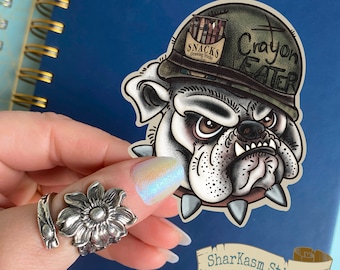 Może przedstawiać: Biały buldog w zielonym hełmie wojskowym z napisem "Crayon Eater" i "Snacks Drawing Stuff". Buldog ma poważny wyraz twarzy i nosi kolczasty kołnierz. Naklejka znajduje się na niebieskim tle.