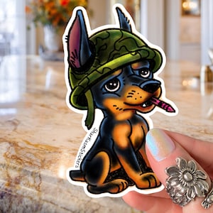 May include: A sticker featuring a cartoon Doberman Pinscher wearing a green camouflage helmet and holding a pink crayon. The dog is black and tan with blue ears and a sad expression. The sticker has the text "SharkasmStickers".