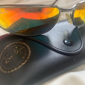 May include: Silver aviator sunglasses with orange mirrored lenses and a black leather case with the Ray-Ban logo.