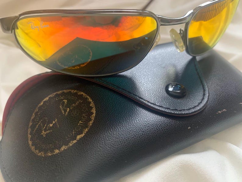 May include: Silver aviator sunglasses with orange mirrored lenses and a black leather case with the Ray-Ban logo.
