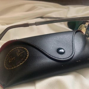 May include: A black leather case with a gold Ray-Ban logo and a silver and green pair of sunglasses.