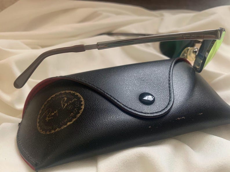 May include: A black leather case with a gold Ray-Ban logo and a silver and green pair of sunglasses.