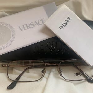 May include: A pair of brown metal eyeglasses with a rectangular frame. The glasses are resting on a black case with the word "VERSUS VERSACE" embossed on it. The case is open and there are two white cards with the word "VERSACE" printed on them.