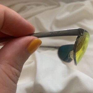 May include: Close-up of a pair of silver Ray-Ban sunglasses with green and yellow lenses.