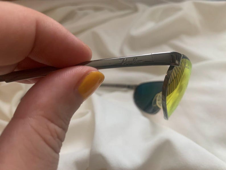 May include: Close-up of a pair of silver Ray-Ban sunglasses with green and yellow lenses.