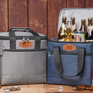 Könnte beinhalten: Zwei isolierte Kühltaschen, eine graue und eine blaue, mit schwarzen Griffen und personalisierten Lederaufnähern. Die blaue Kühltasche ist geöffnet und zeigt Bierflaschen und eine Dose. Die graue Kühltasche trägt den Namen "Bentley".