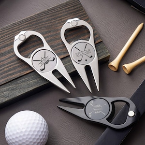 Könnte beinhalten: Drei Golf-Pitchgabeln aus Metall mit integrierten Flaschenöffnern. Zwei sind silberfarben, eine ist schwarz. Jede hat eine kreisförmige Mitte mit eingravierten gekreuzten Golfschlägern und den Namen Wilbert, Steward oder RUA. Enthält zwei Golf-Tees aus Holz.