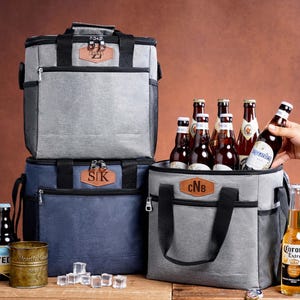 May include: Three insulated cooler bags in gray and navy blue, each featuring a brown leather-like patch with personalized initials. The bags have black handles, zippers, and side mesh pockets. Several glass beer bottles and ice cubes are present.