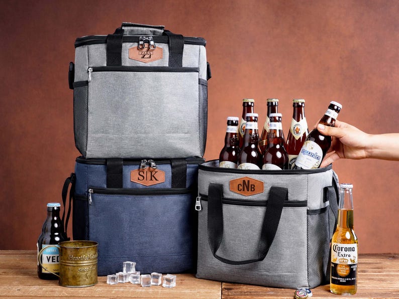 May include: Three insulated cooler bags in gray and navy blue, each featuring a brown leather-like patch with personalized initials. The bags have black handles, zippers, and side mesh pockets. Several glass beer bottles and ice cubes are present.