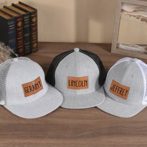Puede incluir: Tres gorras de béisbol grises y blancas con parches de cuero marrón. Los parches tienen los nombres "Geraint", "Lincoln" y "Jeffrey" escritos en ellos.