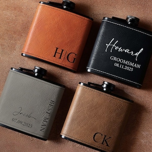 May include: Four leather flasks in shades of brown, black, and gray, each with a black cap. The flasks are personalized with initials, names, and dates. The flasks are arranged on a brown surface.