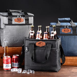 May include: Three insulated cooler bags in grey, black, and blue, each with a handle and shoulder strap. The bags have personalised leather patches and are filled with beer bottles and cans. Ice cubes are scattered in front.