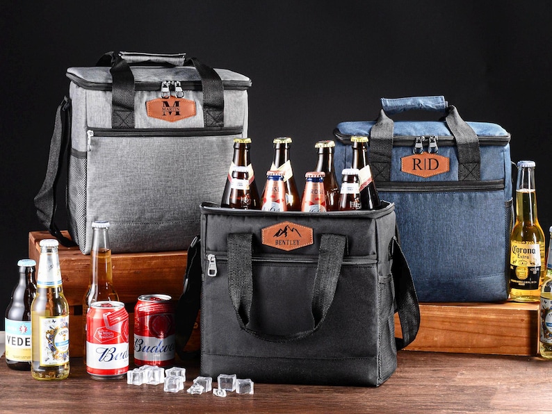 May include: Three insulated cooler bags in gray, black, and blue, each featuring a brown leather-like patch with personalized text. The bags have black handles, zipper closures, and side mesh pockets. Several glass beer bottles and soda cans are present.