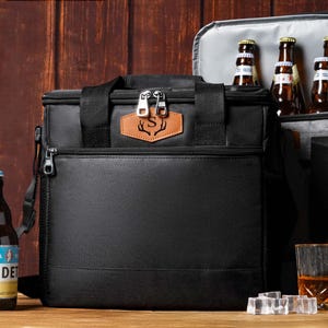 May include: A black insulated cooler bag with a tan leather patch featuring a letter S and antler design. The bag has a top handle, a front zippered pocket, and two silver zipper pulls. An open grey cooler bag sits behind it containing glass bottles.