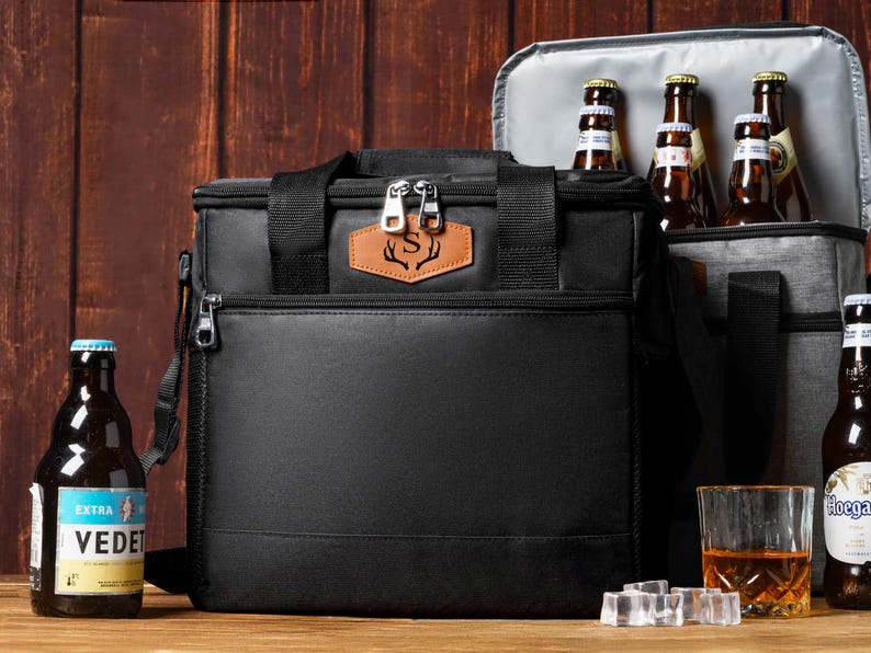 May include: A black insulated cooler bag with a tan leather patch featuring a letter S and antler design. The bag has a top handle, a front zippered pocket, and two silver zipper pulls. An open grey cooler bag sits behind it containing glass bottles.