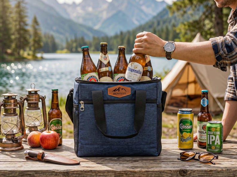 May include: A navy blue insulated cooler bag with a brown leather-style patch labeled Bentley, filled with various glass beer bottles. The bag features black handles, a front zipper pocket, and is placed on a wooden table with beer bottles, cans, apples, lanterns, and sunglasses.
