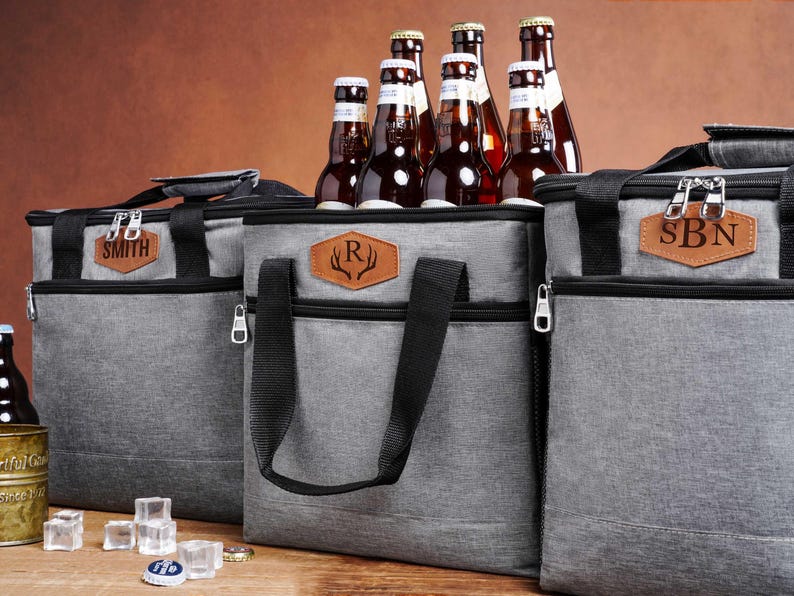 May include: Three grey insulated cooler bags with black straps and zippers. Each bag features a brown leather-like patch with personalized text: SMITH, an R with antlers, and SBN. Several brown glass beer bottles are tucked inside the bags.