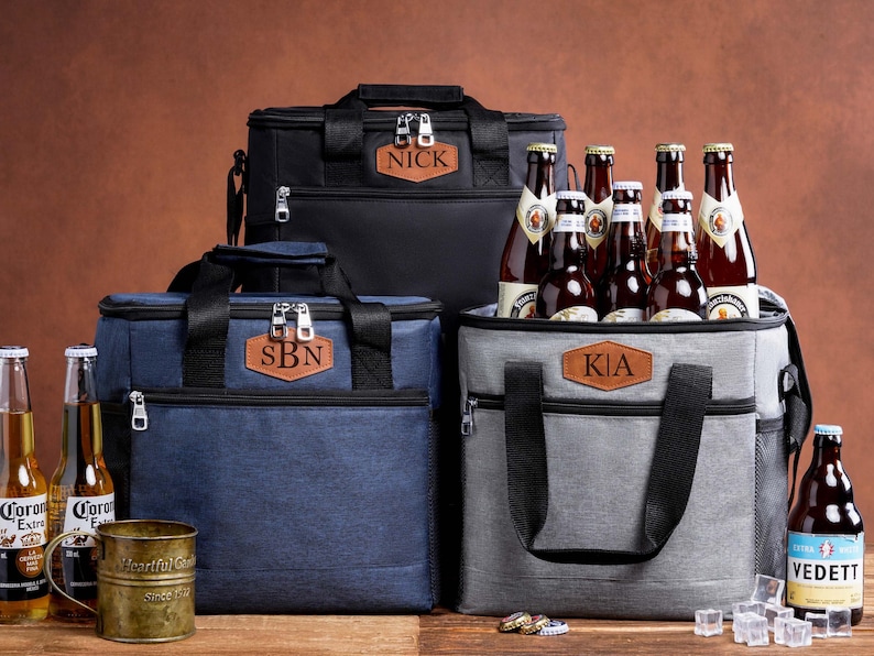 May include: Three insulated cooler bags in black, navy blue, and gray, each featuring a brown leather patch with personalized text: NICK, SBN, and KIA. The bags have black handles, zippers, and side pockets, and are surrounded by beer bottles.