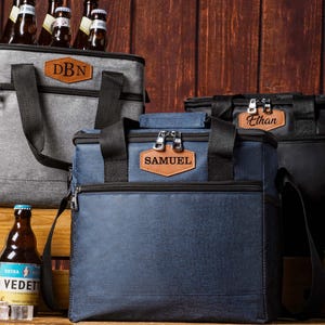 May include: Three insulated cooler bags in gray, navy blue, and black, each featuring a brown leather-like patch with personalized names or initials including DBN, SAMUEL, and Ethan, equipped with black straps and zippers for carrying.
