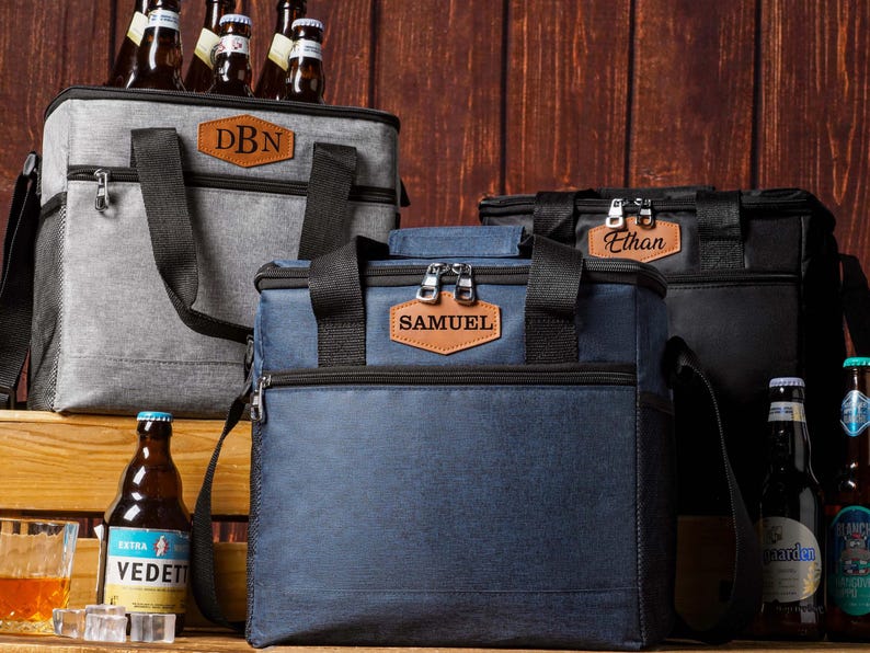 May include: Three insulated cooler bags in gray, navy blue, and black, each featuring a brown leather-like patch with personalized names or initials including DBN, SAMUEL, and Ethan, equipped with black straps and zippers for carrying.