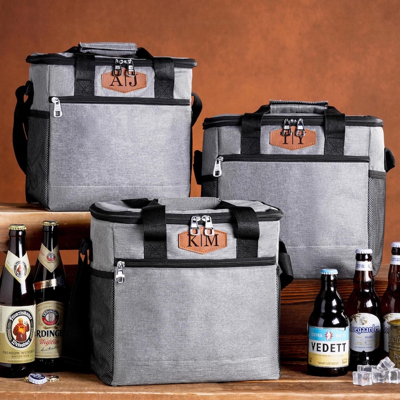 May include: Three grey insulated cooler bags with black trim and handles. Each bag features a brown leather-like patch with personalized initials: AJ, TI, and KIM. The bags have zippered compartments, side mesh pockets, and are displayed with beer bottles.