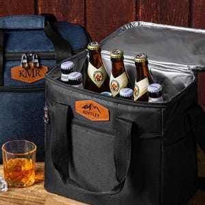 May include: A black insulated cooler bag with a tan leather patch featuring the text BENTLEY and a mountain graphic. The bag is open, revealing several glass beer bottles inside. A second blue cooler bag with a KMR patch sits in the background.