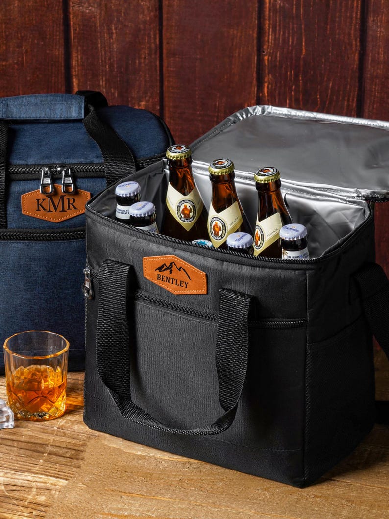 May include: A black insulated cooler bag with a tan leather patch featuring the text BENTLEY and a mountain graphic. The bag is open, revealing several glass beer bottles inside. A second blue cooler bag with a KMR patch sits in the background.