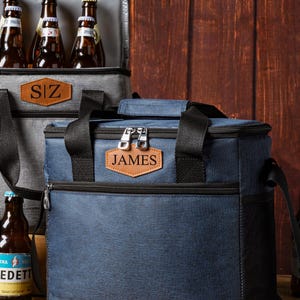 May include: A navy blue insulated cooler bag with black straps, a zippered top, and a brown leather-like patch embossed with the name JAMES. A second grey cooler bag is visible behind it, containing several brown glass beer bottles.