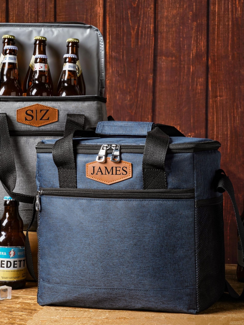 May include: A navy blue insulated cooler bag with black straps, a zippered top, and a brown leather-like patch embossed with the name JAMES. A second grey cooler bag is visible behind it, containing several brown glass beer bottles.