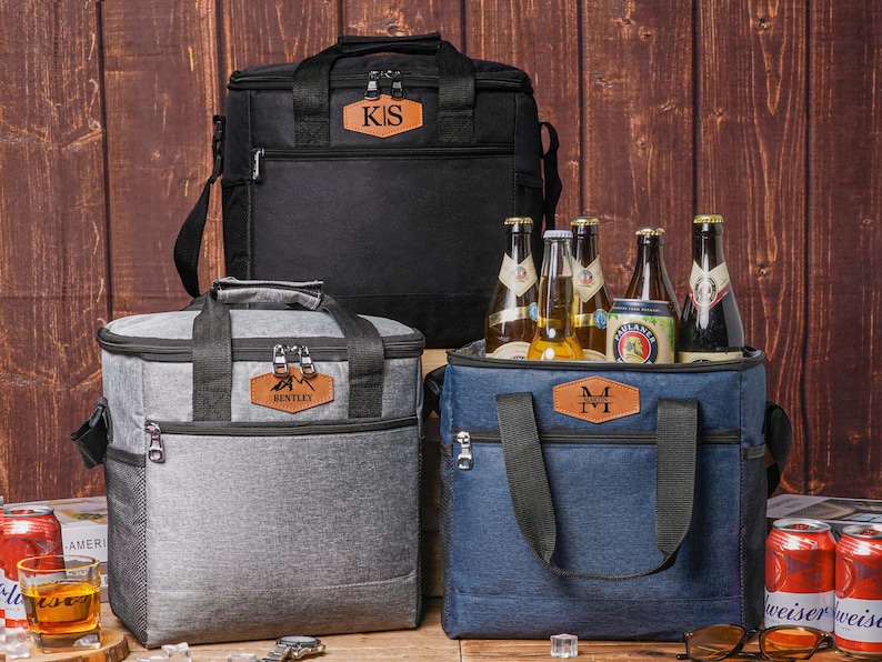 May include: Three insulated cooler bags in black, gray, and blue, each featuring a rectangular leather patch with personalized initials or names. The bags have black handles, zippered closures, and side mesh pockets. One bag is filled with glass bottles.