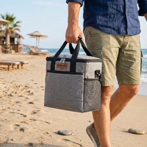 May include: A rectangular gray insulated cooler bag with black trim, black carrying handles, and a side mesh pocket. A brown leather-like patch on the front features the name MARTIN embossed in black text. The bag has a zippered top closure.