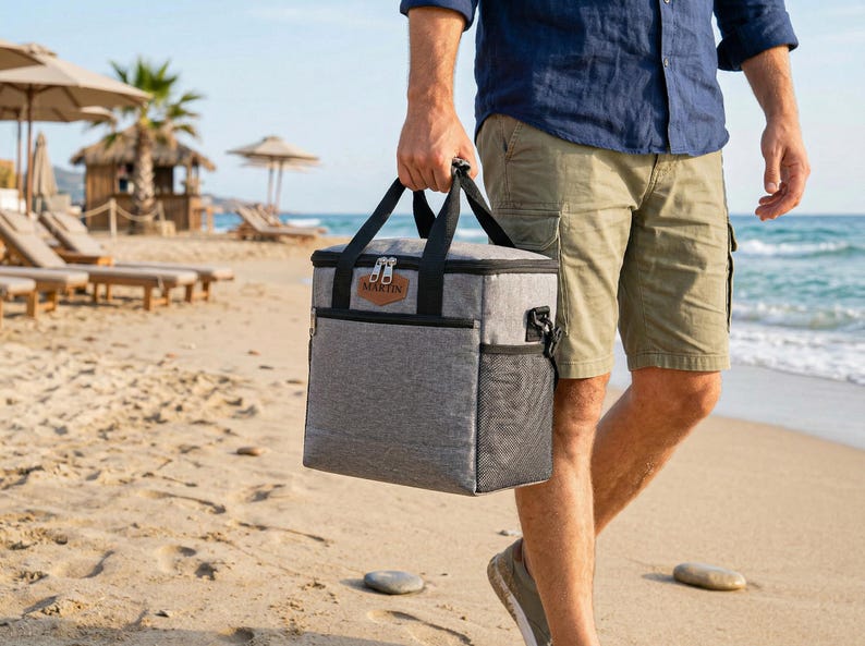 May include: A rectangular gray insulated cooler bag with black trim, black carrying handles, and a side mesh pocket. A brown leather-like patch on the front features the name MARTIN embossed in black text. The bag has a zippered top closure.