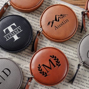 Puede incluir: Una colección de cintas métricas redondas personalizadas en varios colores, incluyendo negro, marrón y gris. Cada cinta métrica presenta texto o un diseño personalizado, como un nombre o iniciales, en blanco o negro. Tienen una correa de cuero.