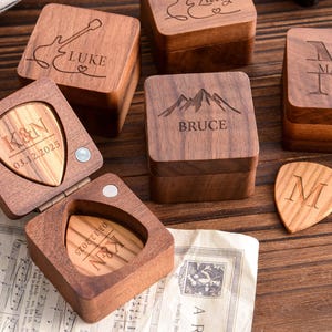 Può includere: Scatole in legno per plettri per chitarra con incisioni personalizzate. Una scatola è aperta, rivelando un plettro con inciso "K&N 03.12.2025". Altre scatole presentano nomi come "LUKE" e "BRUCE", e iniziali.