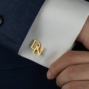May include: Gold-colored cufflinks with the initials "DN" are displayed on a white dress shirt cuff. The cuff is part of a dark blue suit. The cufflinks are a stylish accessory.