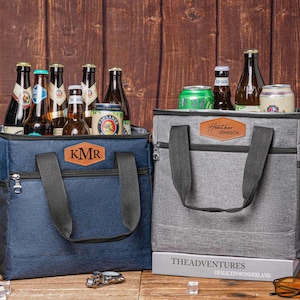 May include: A navy blue cooler bag with a brown leather patch labeled KMR and a grey cooler bag with a brown leather patch labeled Heather Johnson. Both bags feature black handles, front zipper pockets, and side mesh pockets, filled with various glass bottles and aluminum cans.