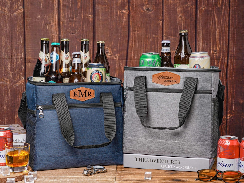 May include: A navy blue cooler bag with a brown leather patch labeled KMR and a grey cooler bag with a brown leather patch labeled Heather Johnson. Both bags feature black handles, front zipper pockets, and side mesh pockets, filled with various glass bottles and aluminum cans.