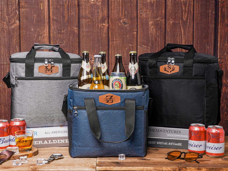 May include: Three insulated cooler bags in gray, blue, and black, each featuring a front zippered pocket and a brown leather-like patch with a personalized letter. The bags have black handles and zippers, and are filled with various glass beer bottles.